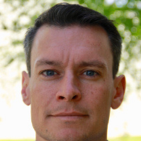 A close-up portrait of a person with short brown hair, blue eyes, and a neutral expression against a blurred green backdrop.