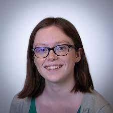 A person with auburn hair and glasses smiling against a plain, light gray background.