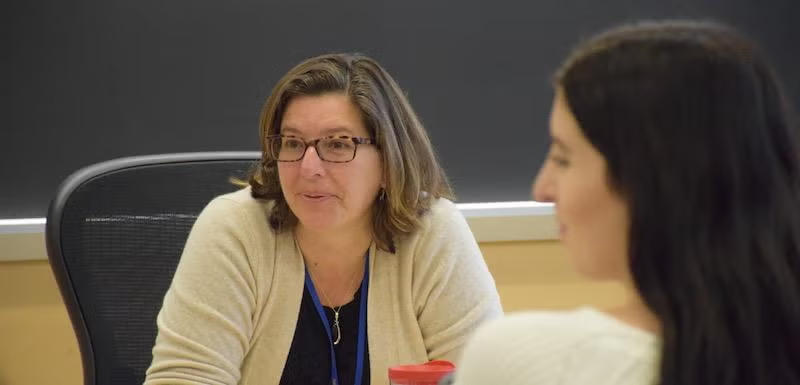 A person with glasses and a cream-colored cardigan sits at a desk during a discussion, facing another person.