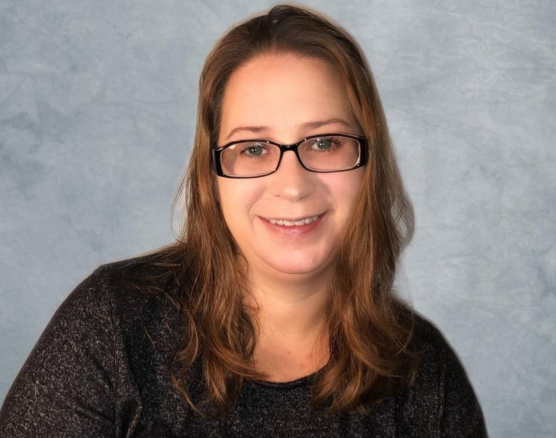 A person with long brown hair and glasses smiles slightly, wearing a dark textured shirt against a mottled blue background.