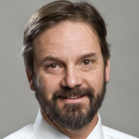A person with short brown hair and a beard, wearing a white collared shirt against a gray background, smiling at the camera.