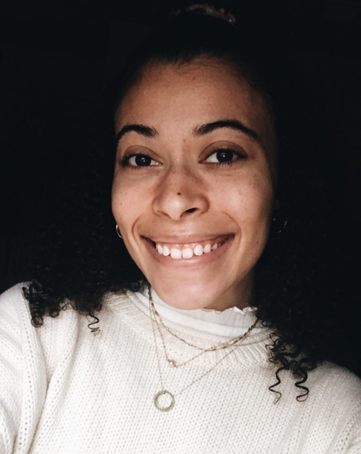 A smiling person with curly dark hair wearing a white textured sweater and layered gold necklaces against a dark background.