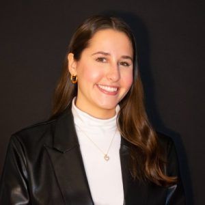 A smiling person wearing a white turtleneck and black blazer against a solid dark background.