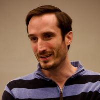 A person with short brown hair and a beard, wearing a blue and dark navy horizontal striped zip-up sweater.