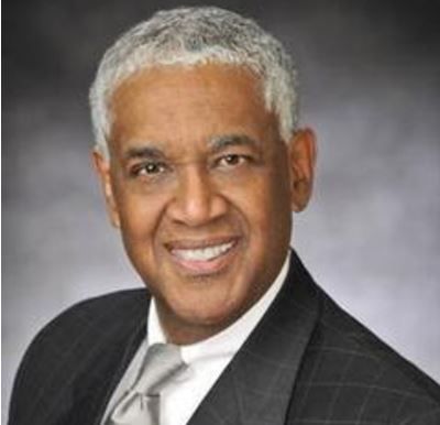 Headshot of a smiling person wearing a dark suit, white shirt, and light grey tie against a neutral grey background.