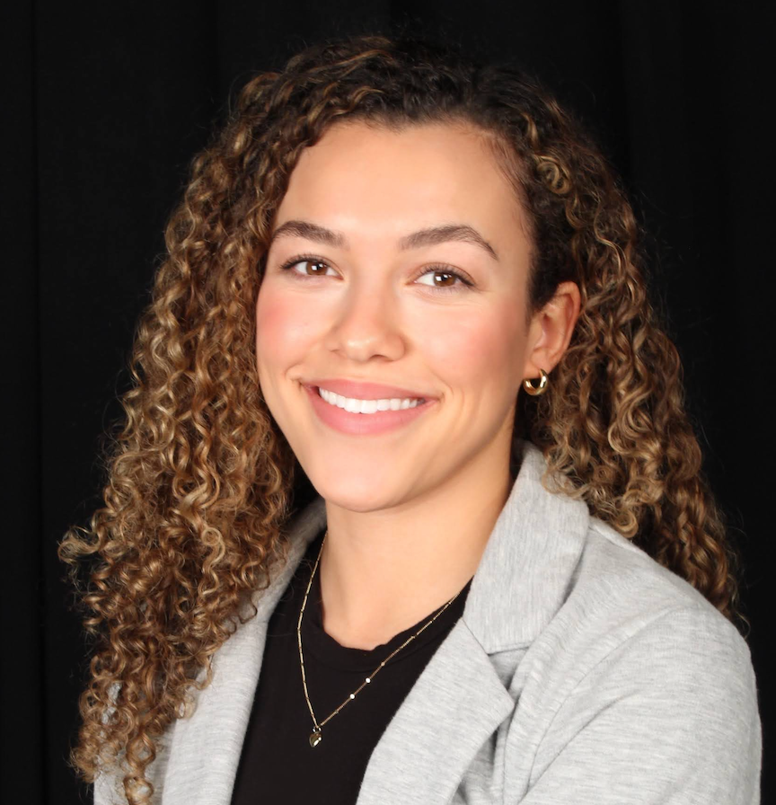Smiling person with curly hair, wearing a gray blazer and gold necklace against a black background.