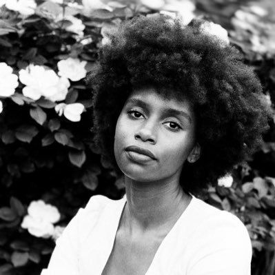 A black-and-white portrait of a person with voluminous curly hair, looking forward in front of a leafy floral background.