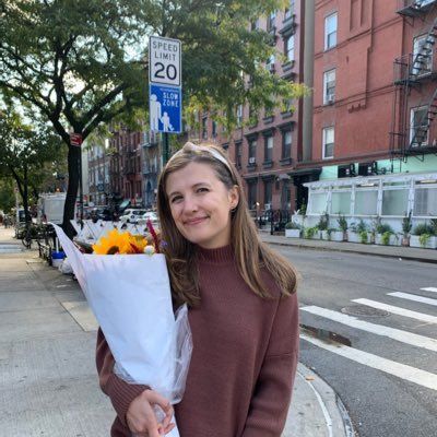 A person in a brown sweater smiles while holding a large bouquet of sunflowers on a city sidewalk next to a speed sign.