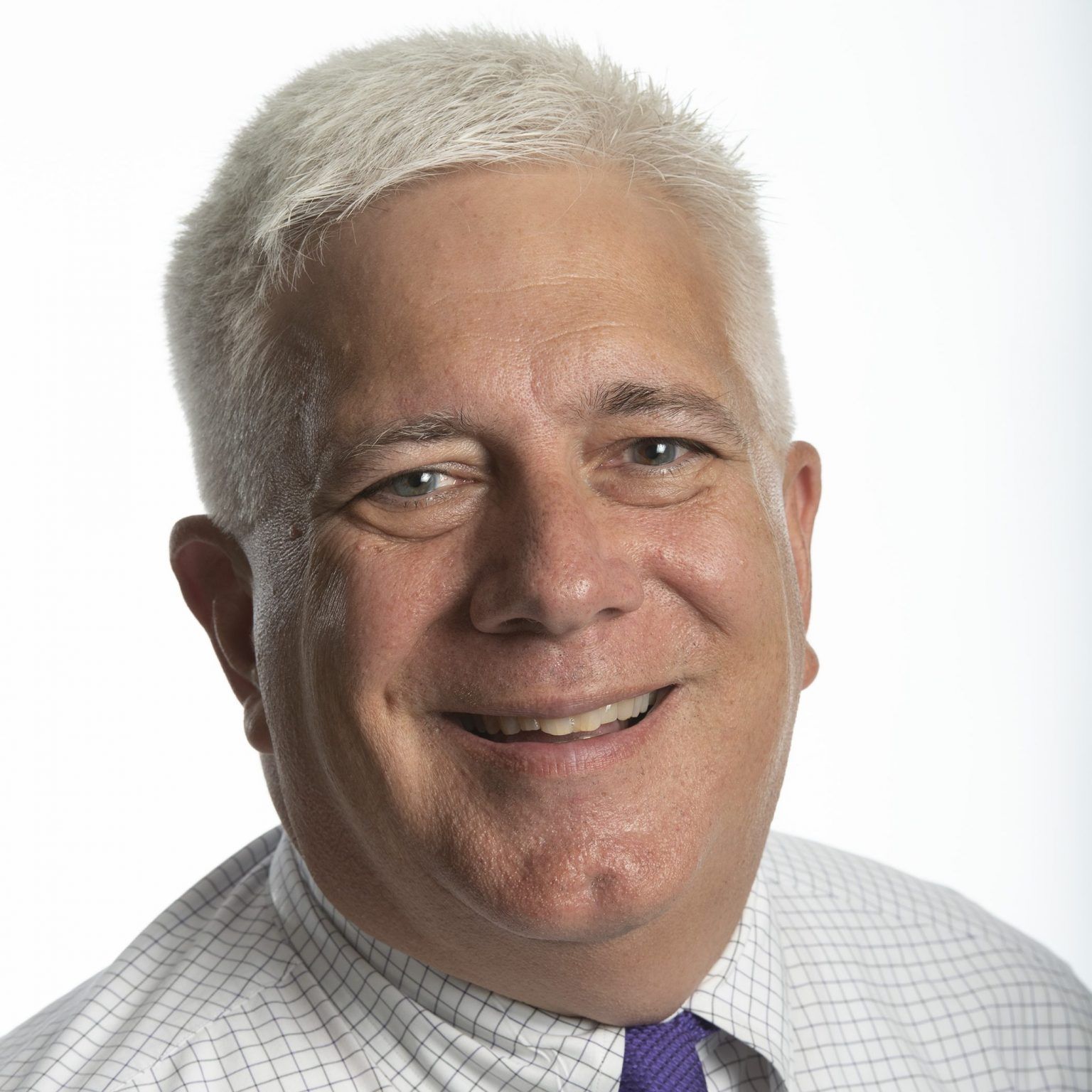 A smiling person with short, white hair, wearing a white checkered collared shirt and a purple tie against a white background.