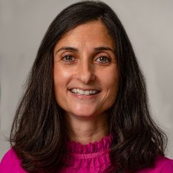 A smiling person with shoulder-length brown hair, wearing a bright pink high-neck top against a plain light gray background.
