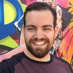 A smiling person with a beard stands in front of a colorful graffiti wall with bright shapes and patterns.