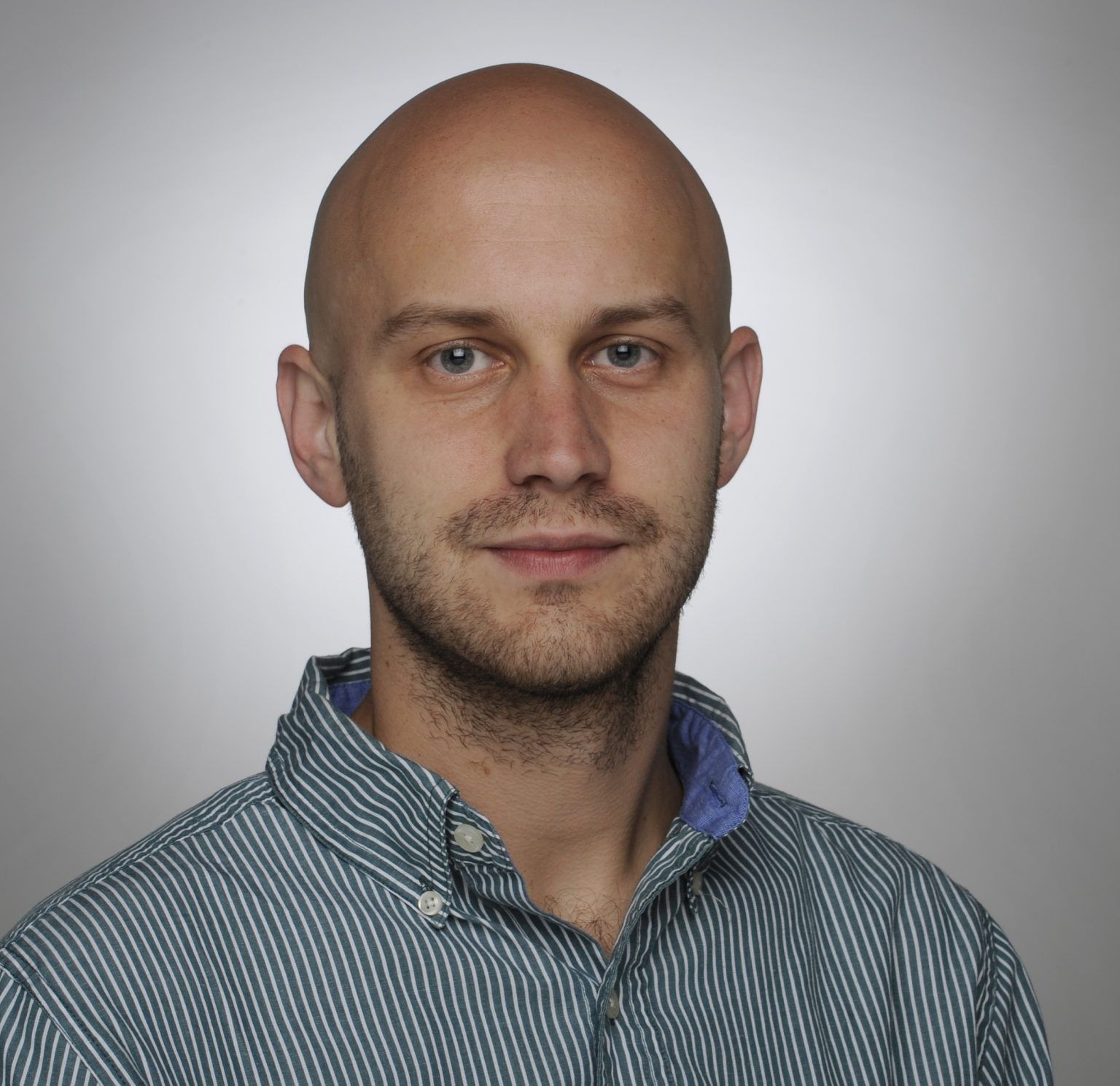 A portrait of a bald person with short facial hair wearing a blue and white striped collared shirt against a gray background.