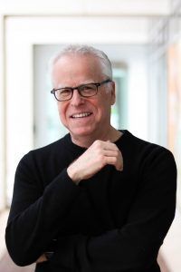 A smiling person with glasses and white hair wearing a black long-sleeved shirt, posed with arms crossed.