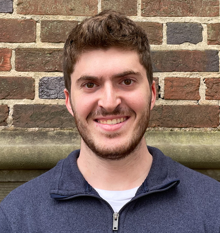 A person with brown hair and facial hair, wearing a blue quarter-zip sweater, smiles in front of a brick wall.