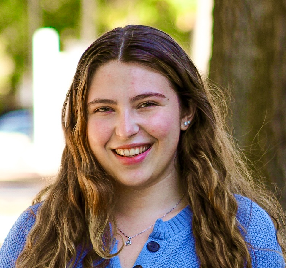 A person with long, wavy brown hair and a bright smile wearing a blue sweater, standing outdoors near a tree.