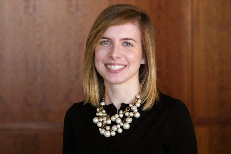 A person with shoulder-length blonde hair wearing a pearl statement necklace, smiling against a wood-paneled background.