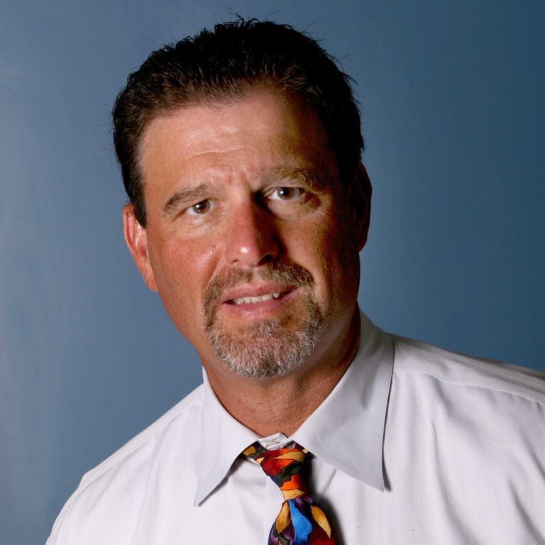 A person with short dark hair and a goatee, wearing a white collared shirt and colorful tie against a blue background.
