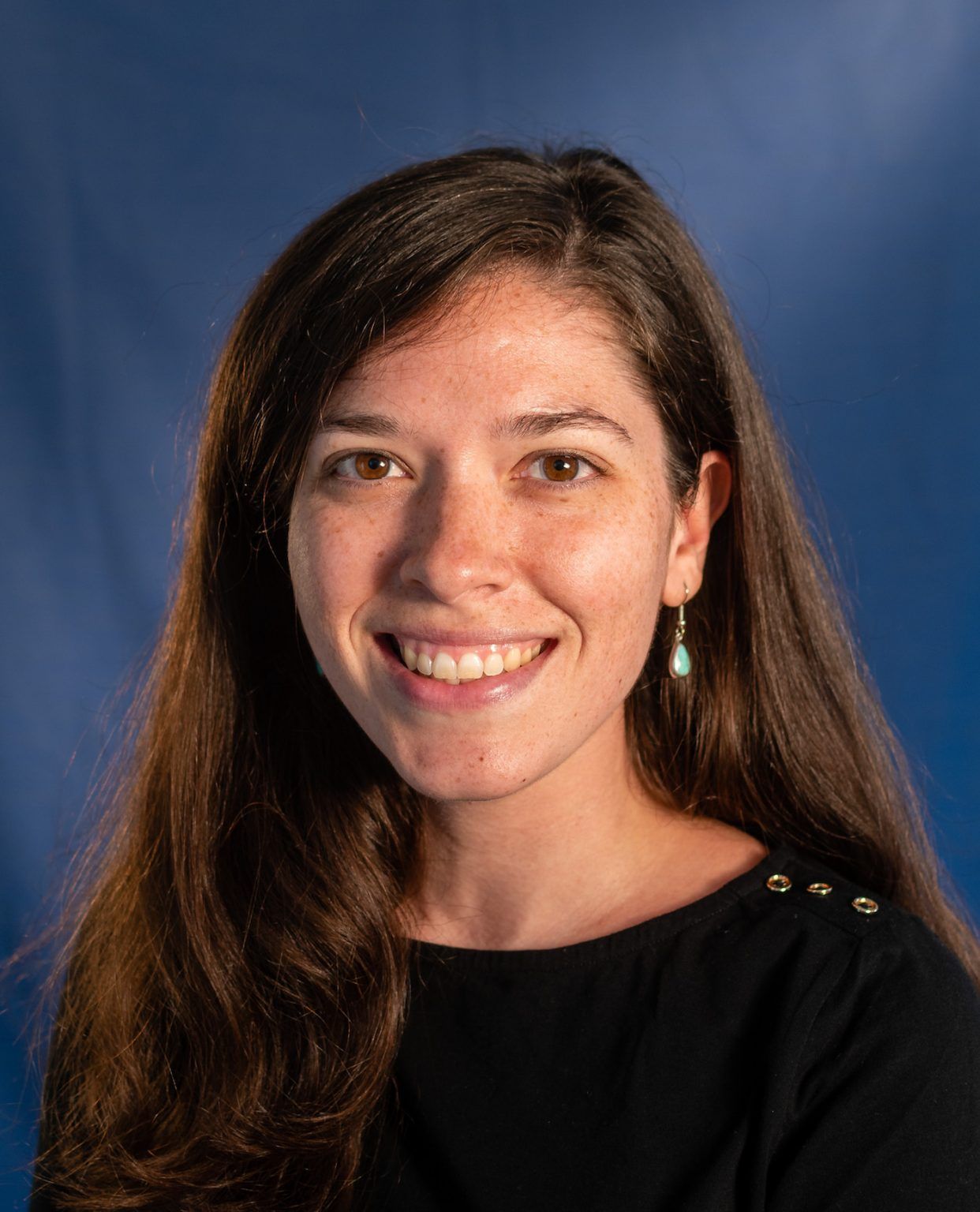 A person with long brown hair smiling against a plain blue background, wearing a black top and small green earrings.