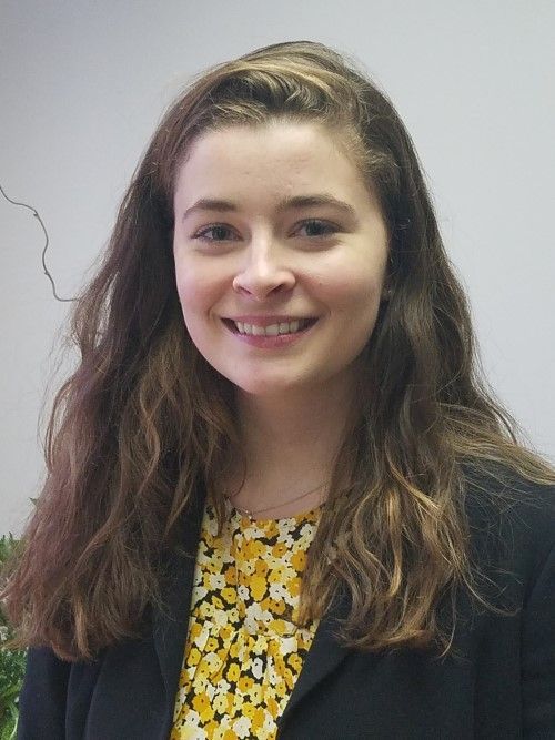 A smiling person with long, wavy brown hair, wearing a black blazer over a yellow and white floral patterned top.