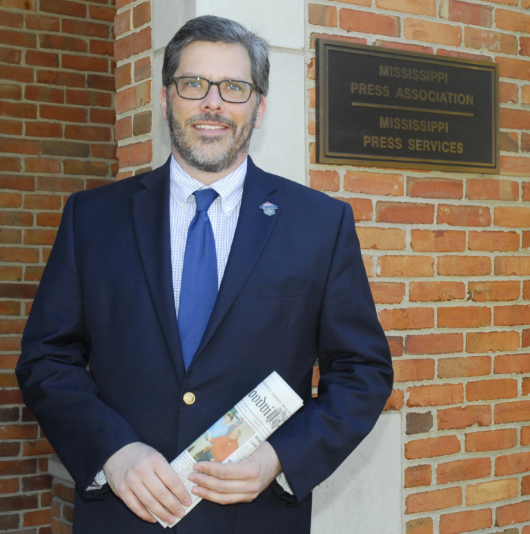 A smiling person in a blue suit and tie holding a newspaper in front of a Mississippi Press Association sign.
