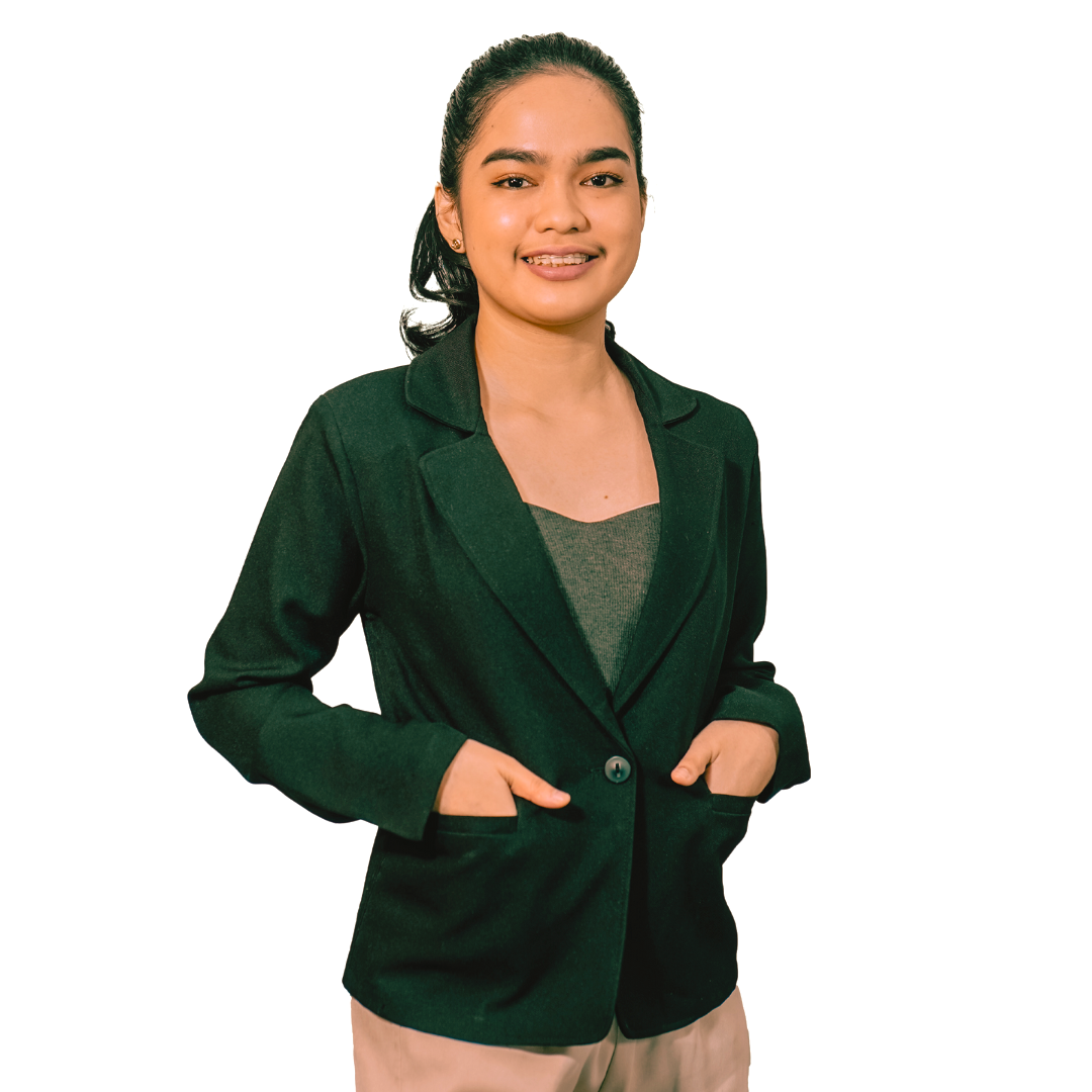 Smiling person with dark hair in a ponytail, wearing a green blazer and grey top, posing with hands in blazer pockets.