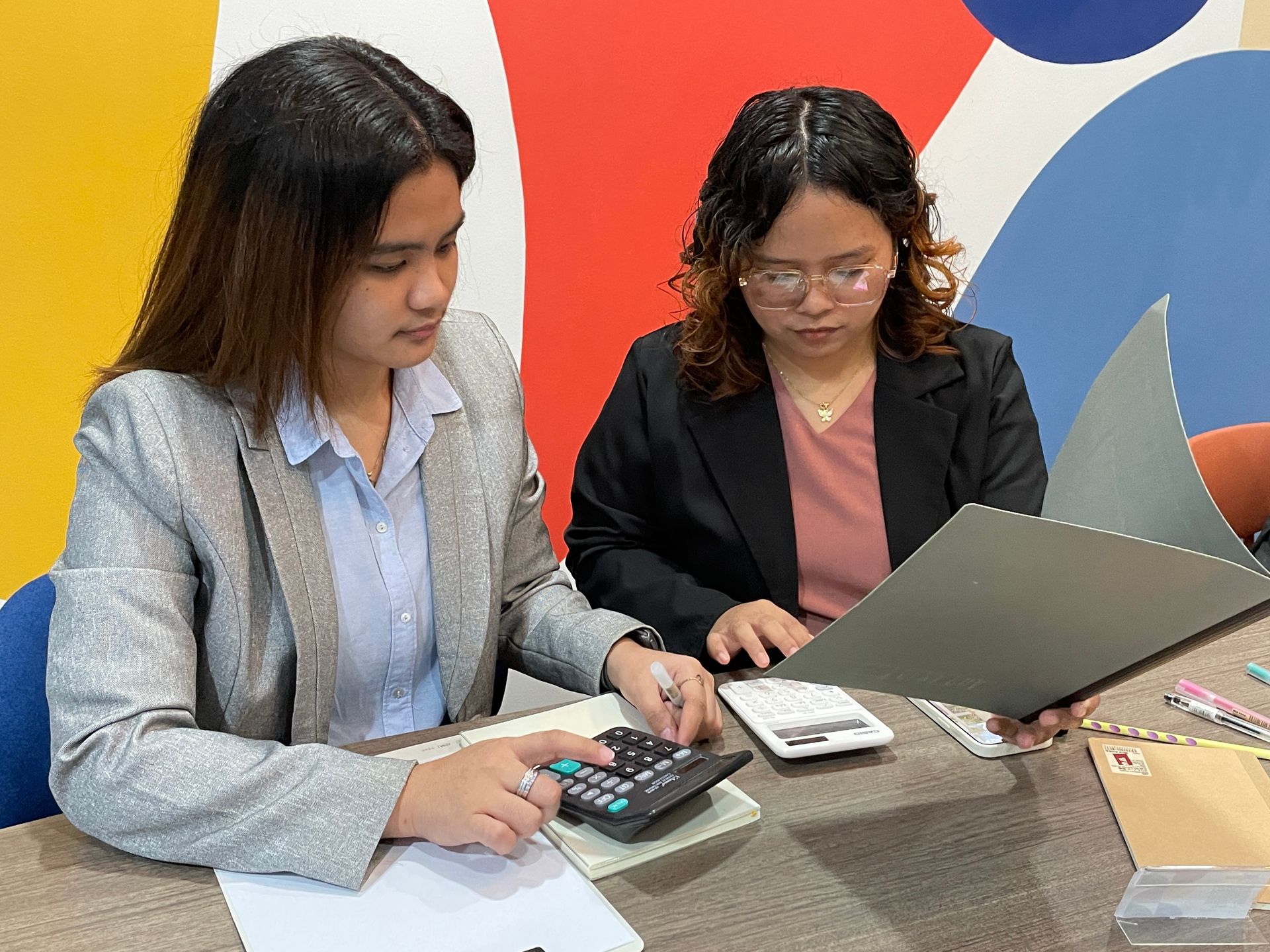Two women are sitting at a table with a calculator and a laptop.