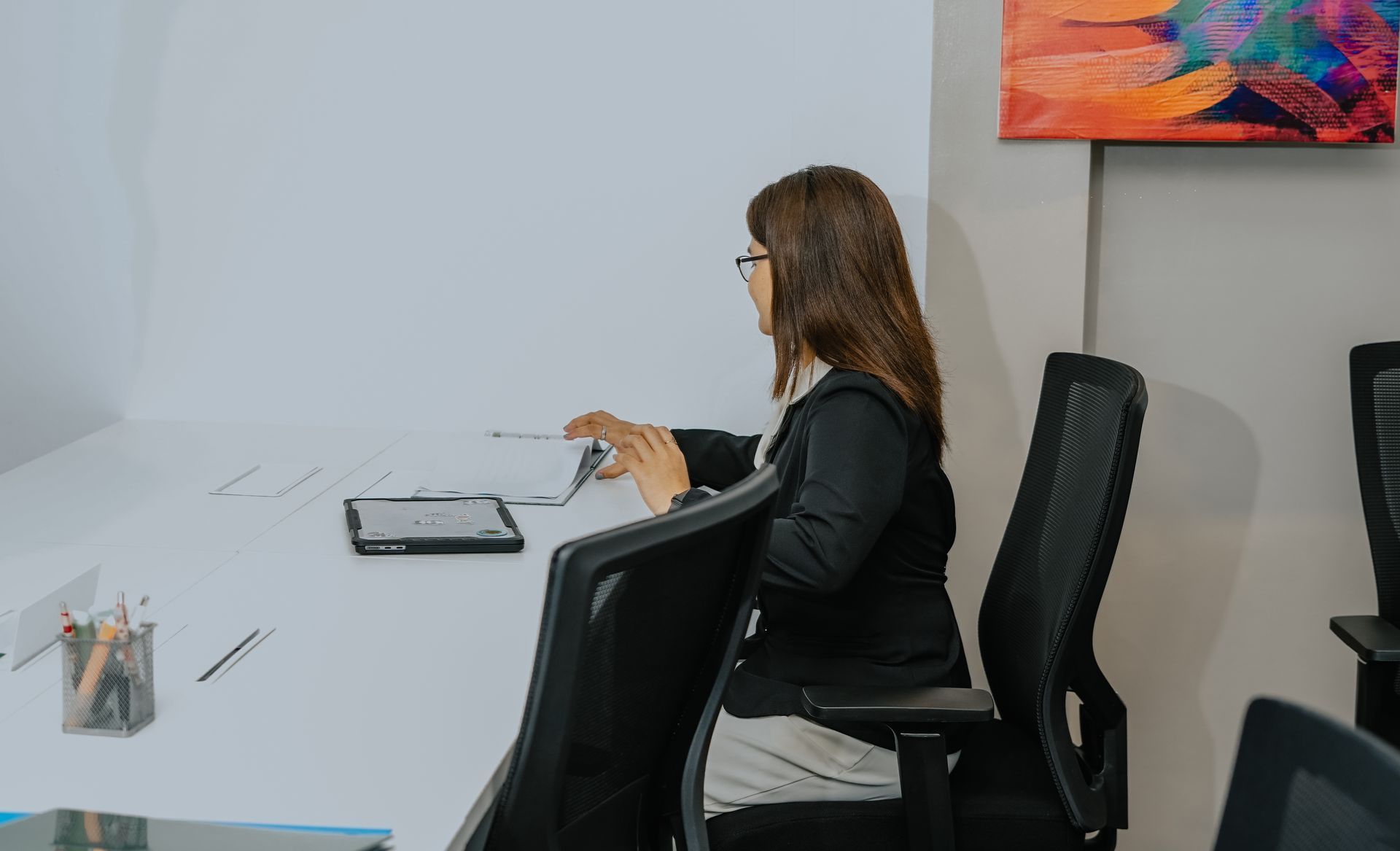 Woman in a black blazer at a white desk, working with papers and a tablet.