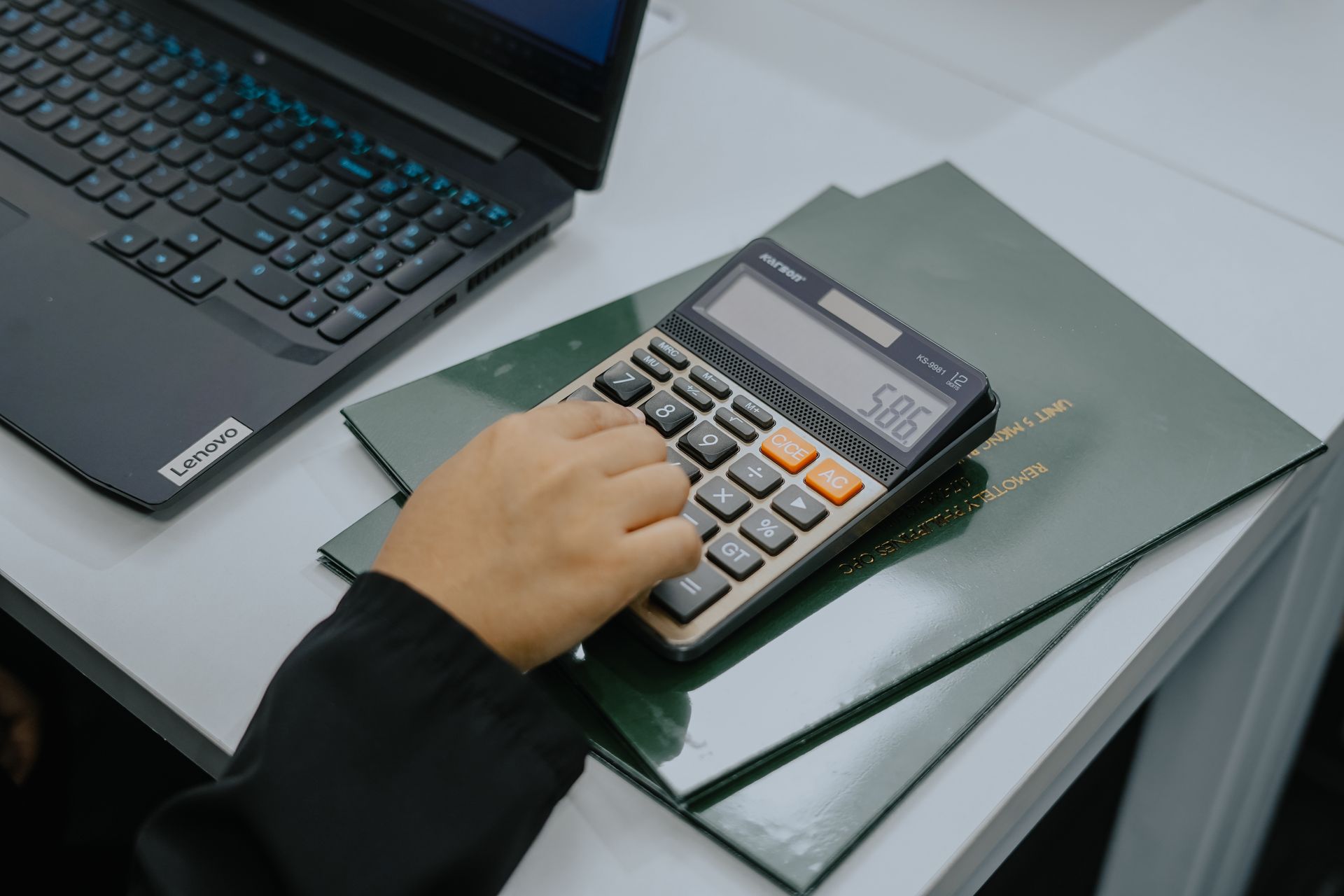 Person using a calculator on top of a stack of green books next to a laptop.