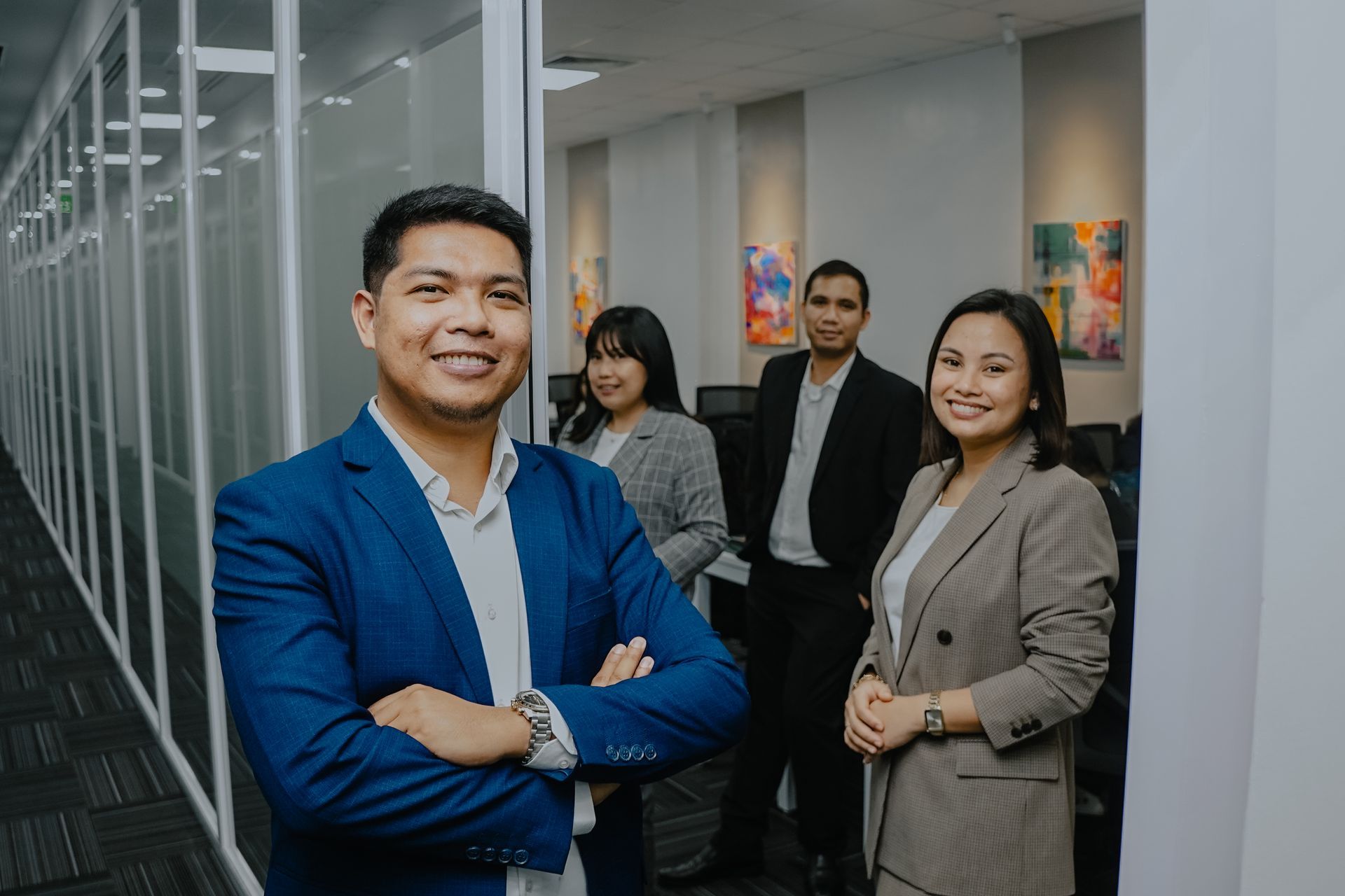 Man in blue blazer smiles, arms crossed, in office. Woman in blazer stands beside him. Other people work in background.