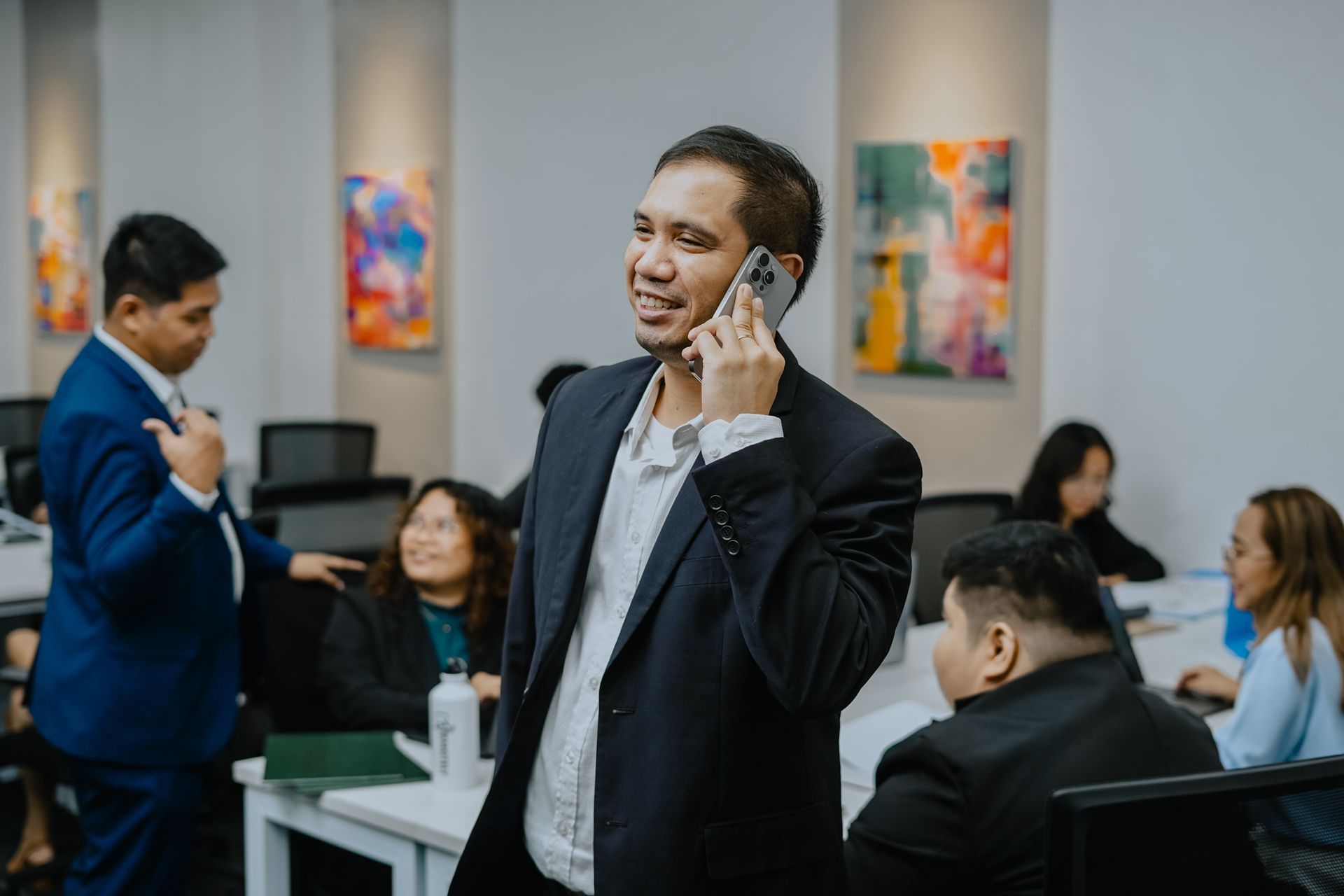 Man in a suit jacket on a phone in an office, smiling. Other people work at desks in the background.