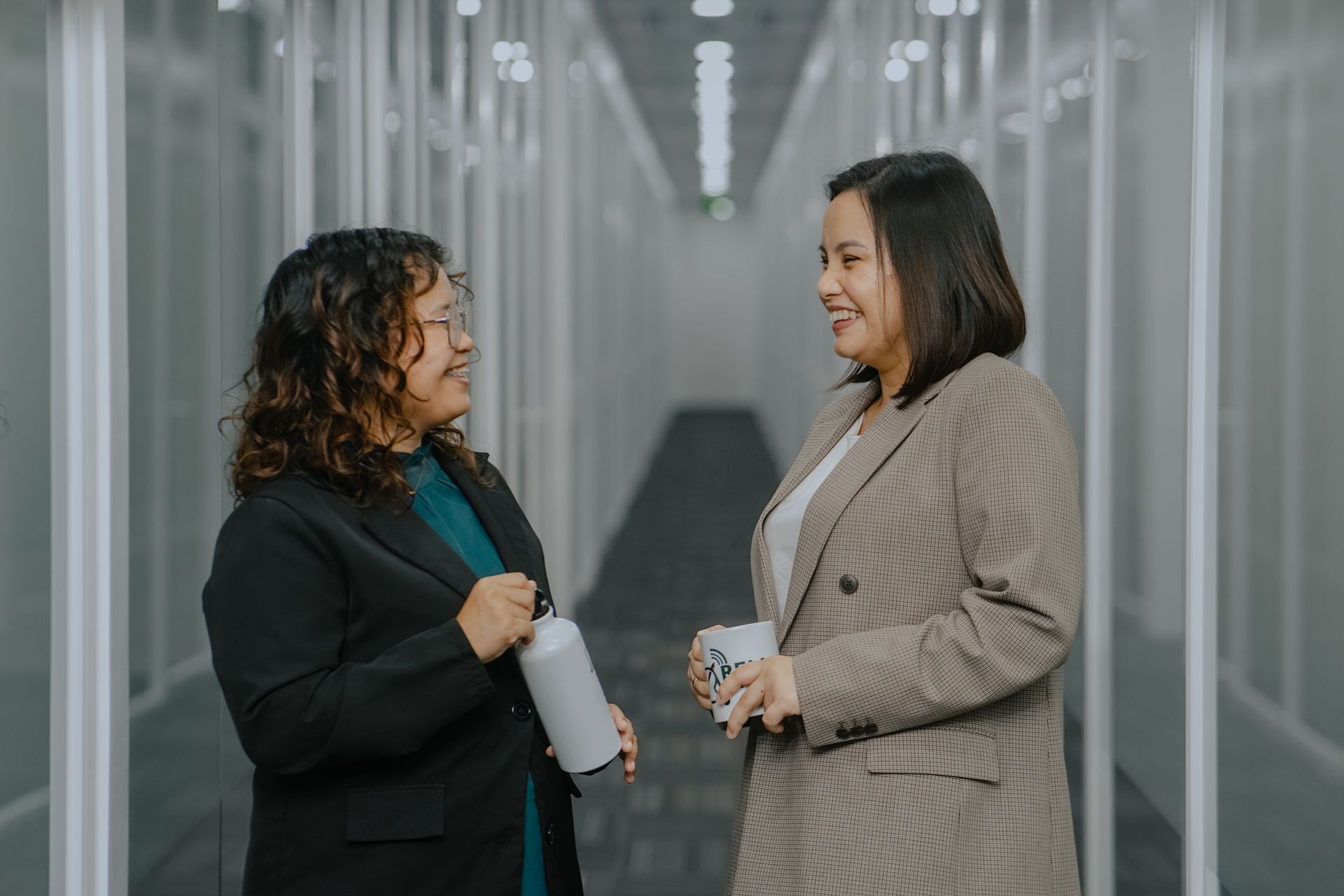 Two professional people talking in a modern hallway with white walls and glass panels, holding drink containers.