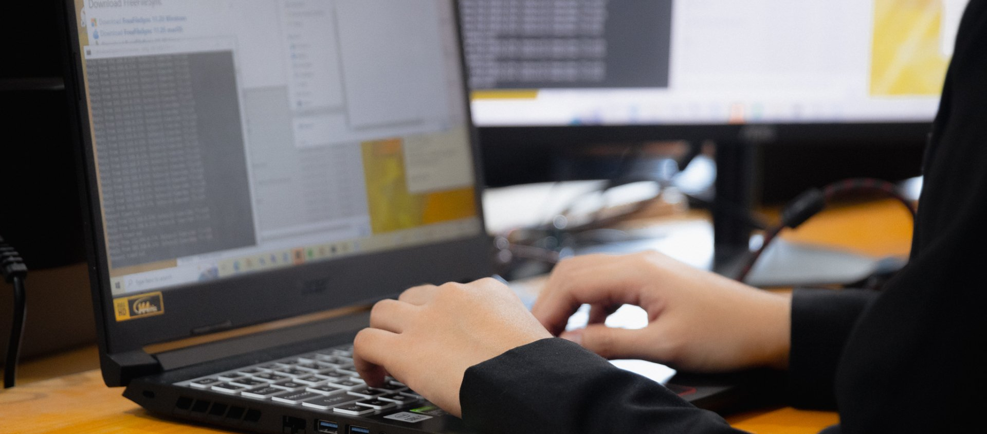 Hands typing on a black laptop keyboard at a desk with an additional computer monitor in the background.