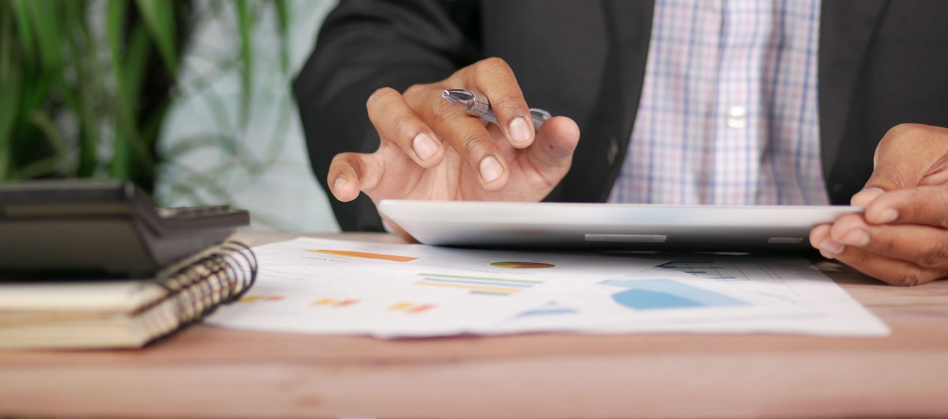 Hands using a tablet on a desk beside a notebook and papers in an office setting