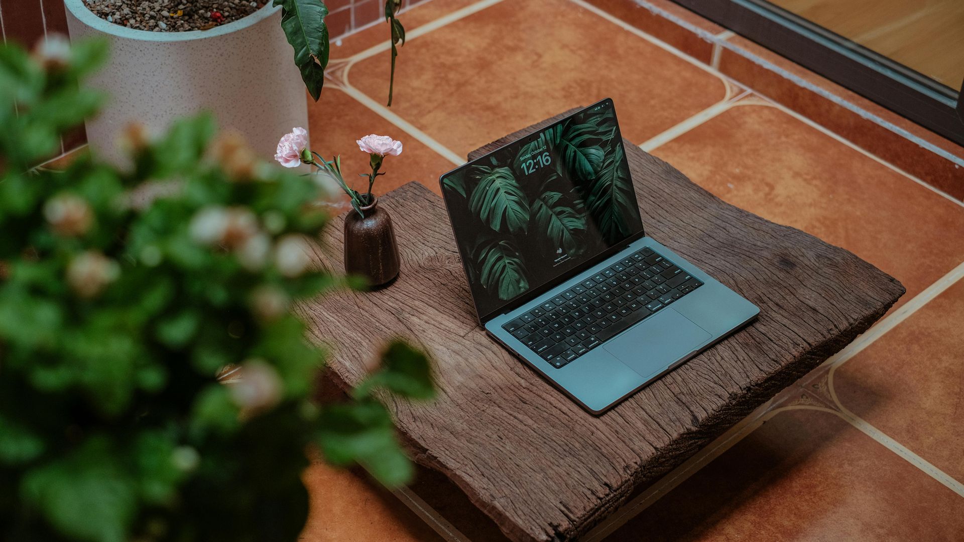 Laptop on a woven mat beside a potted plant, with a small lamp on a tiled floor.