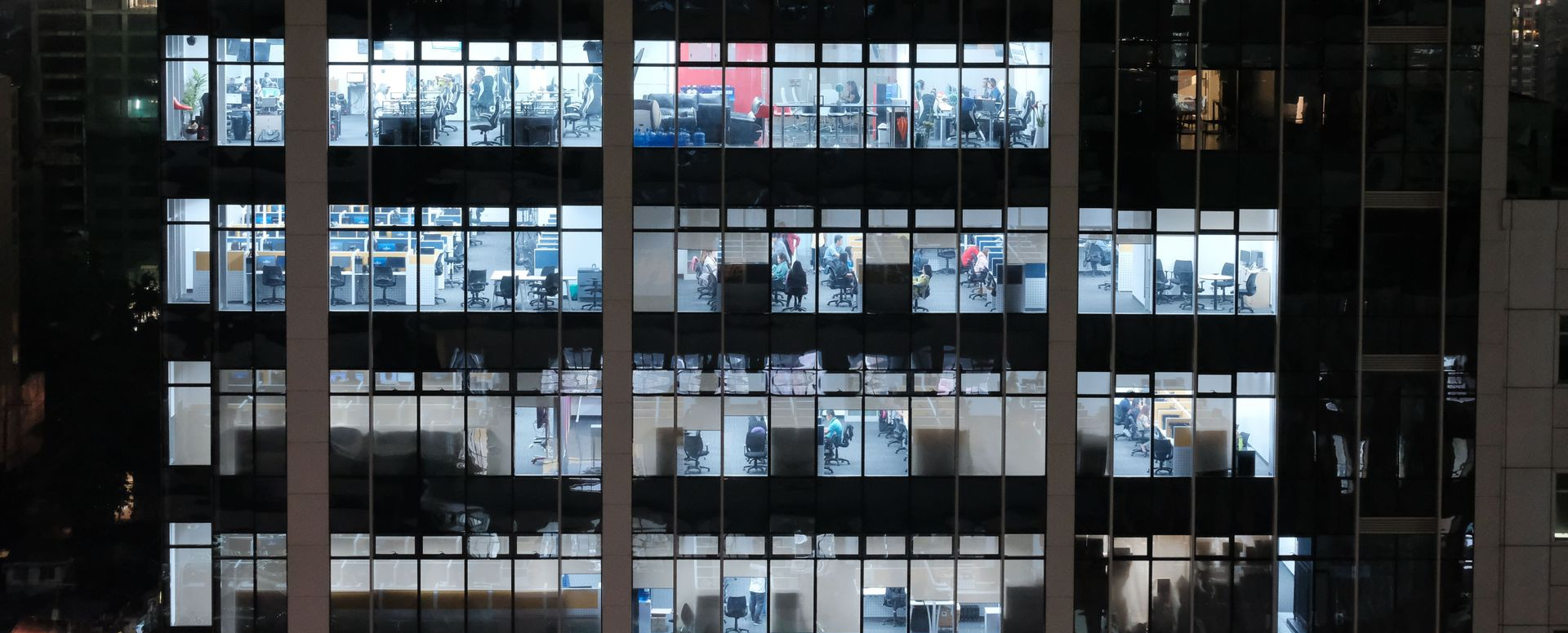 Office building windows glowing at night, with people visible inside across several floors.