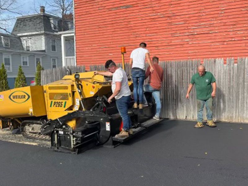 Workers paving asphalt with a yellow paver machine near a red brick building and wooden fence.
