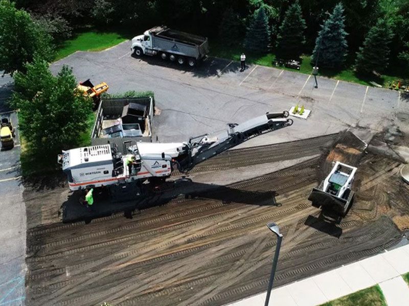 Asphalt milling in progress: Machine grinding pavement, front-end loader, dump truck, and workers on site in parking lot.