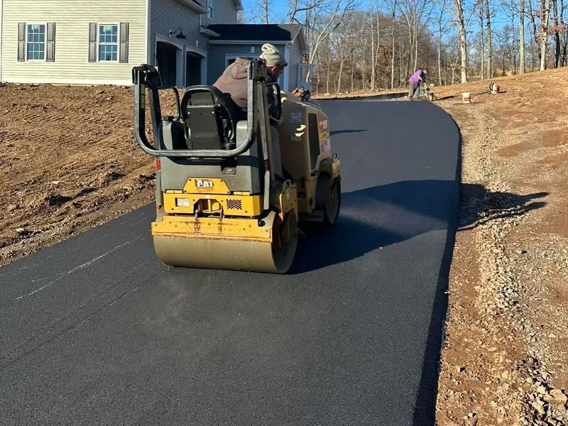 Yellow road roller paving a fresh black driveway beside a house on a sunny day