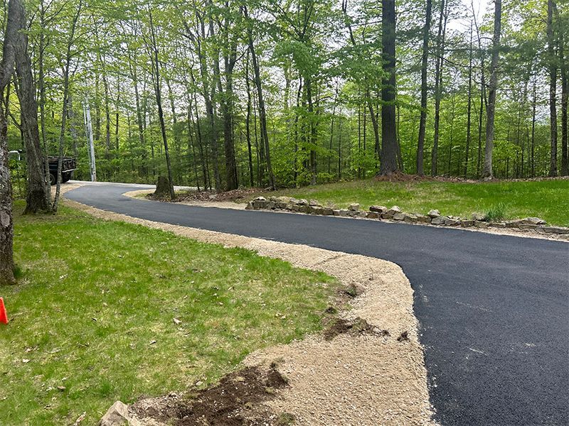 Curving paved path through a wooded park, bordered by grass and gravel.