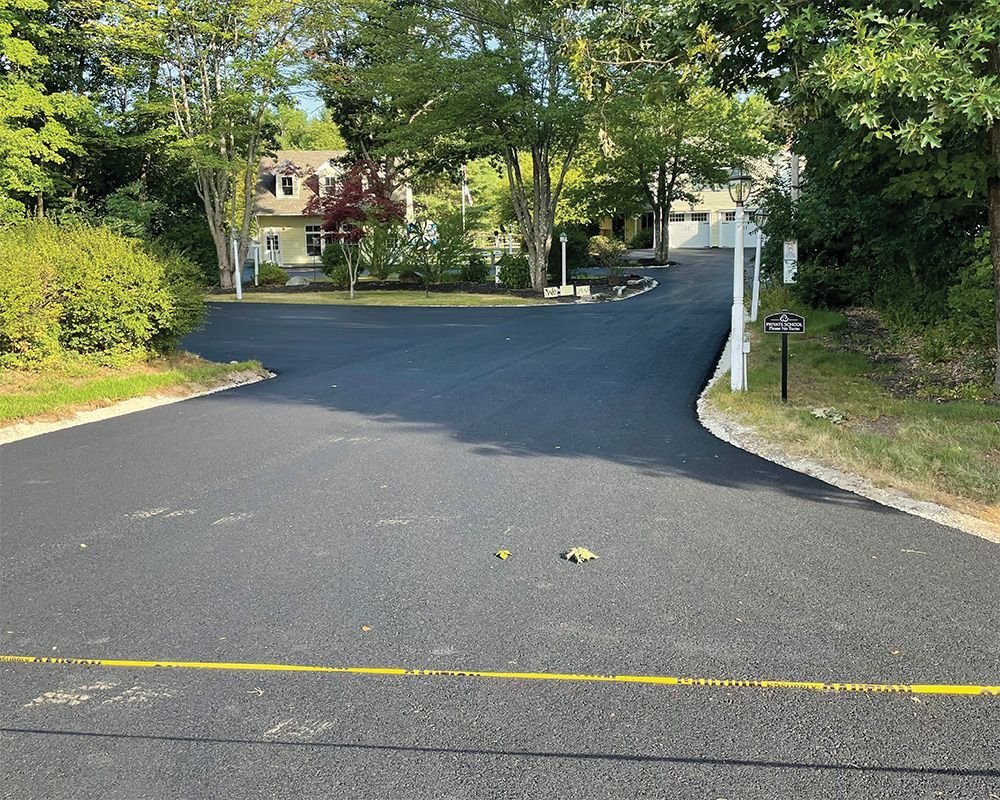 Paved driveway or road entrance bordered by trees and grass, leading to houses in the distance.