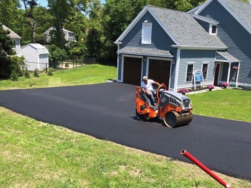 Worker operating an orange asphalt roller on a new driveway beside a gray house.