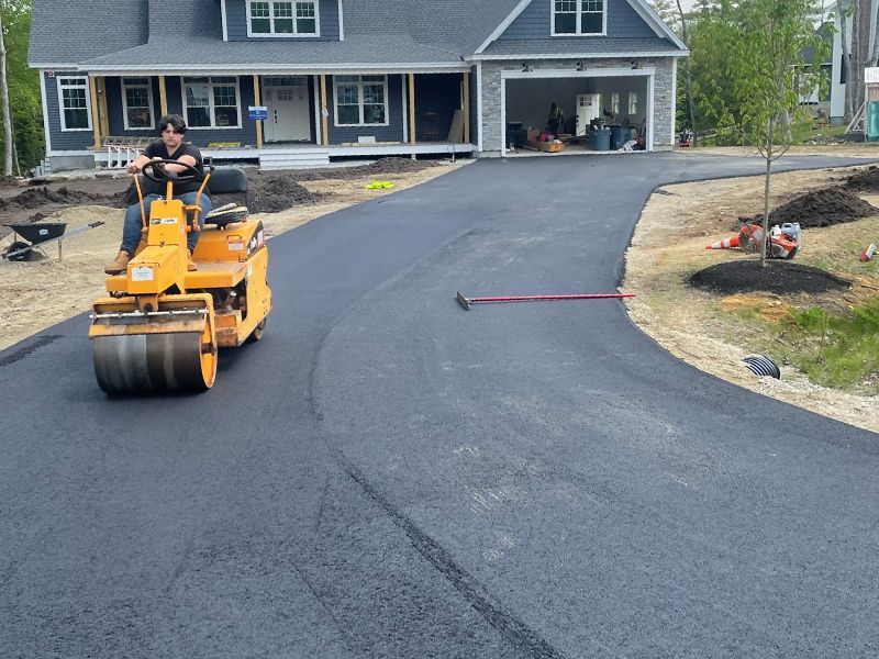Worker on an orange roller paving a fresh black driveway in front of a house