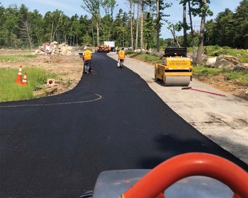 Workers paving a road. Two workers use hand rollers. A roller compactor and construction equipment are visible.