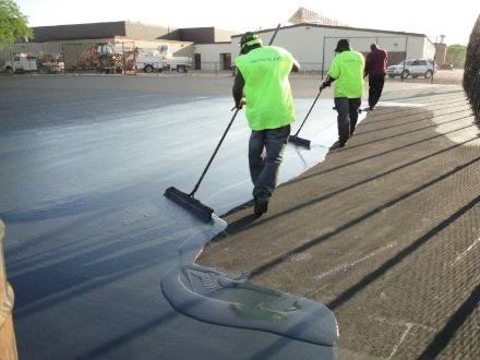 Workers in neon green shirts spread dark sealant on a flat roof with long-handled brooms.