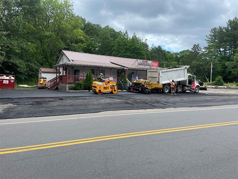 Asphalt paving in progress in front of a building; road roller, paving machine, dump truck, and workers present.