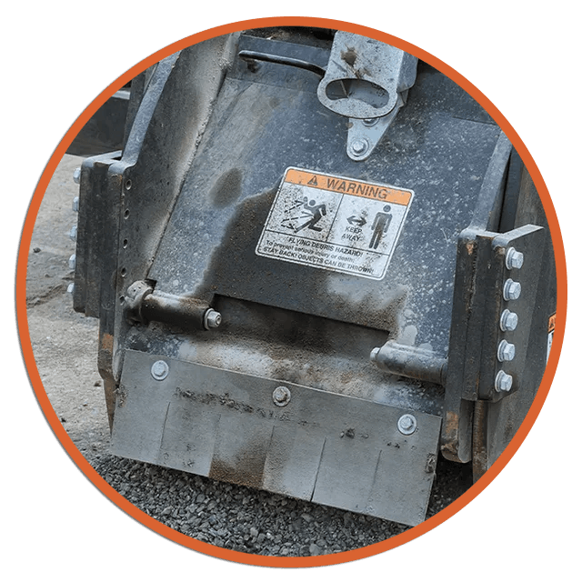 Close-up of a gray metal machine attachment with warning label on gravel, framed by an orange border
