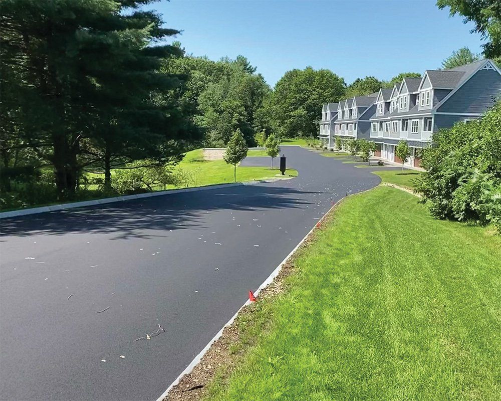 Curving paved road beside green lawn and trees, with townhouses under a clear blue sky.
