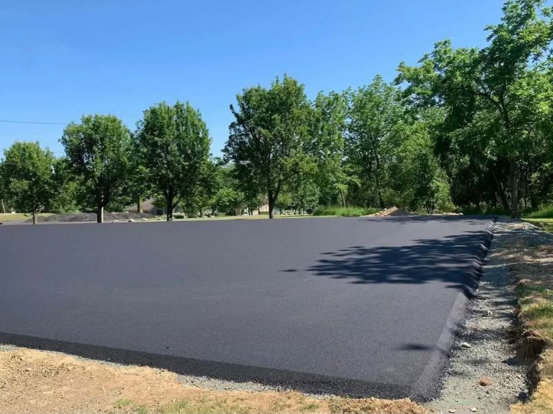 Freshly paved black asphalt lot beside gravel and trees under a clear blue sky