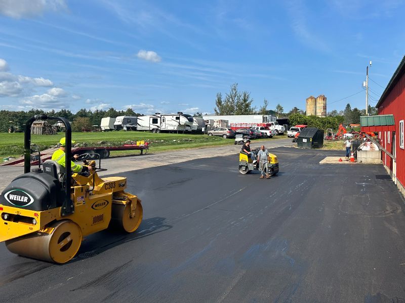 Asphalt paving in progress: two rollers compacting fresh black asphalt surface. Bright yellow equipment and workers are visible.
