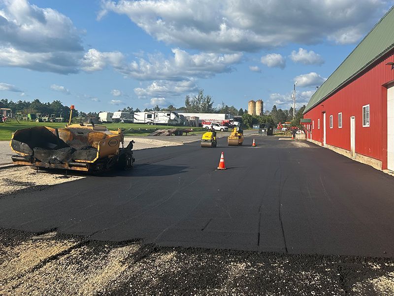 Asphalt paving in progress next to a red building under a cloudy blue sky. A paver and rollers are present.